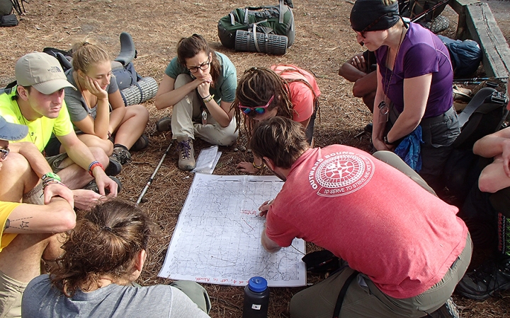 A map lays on the ground and people sit around it. One person, who appears to be an instructor, points at the map.